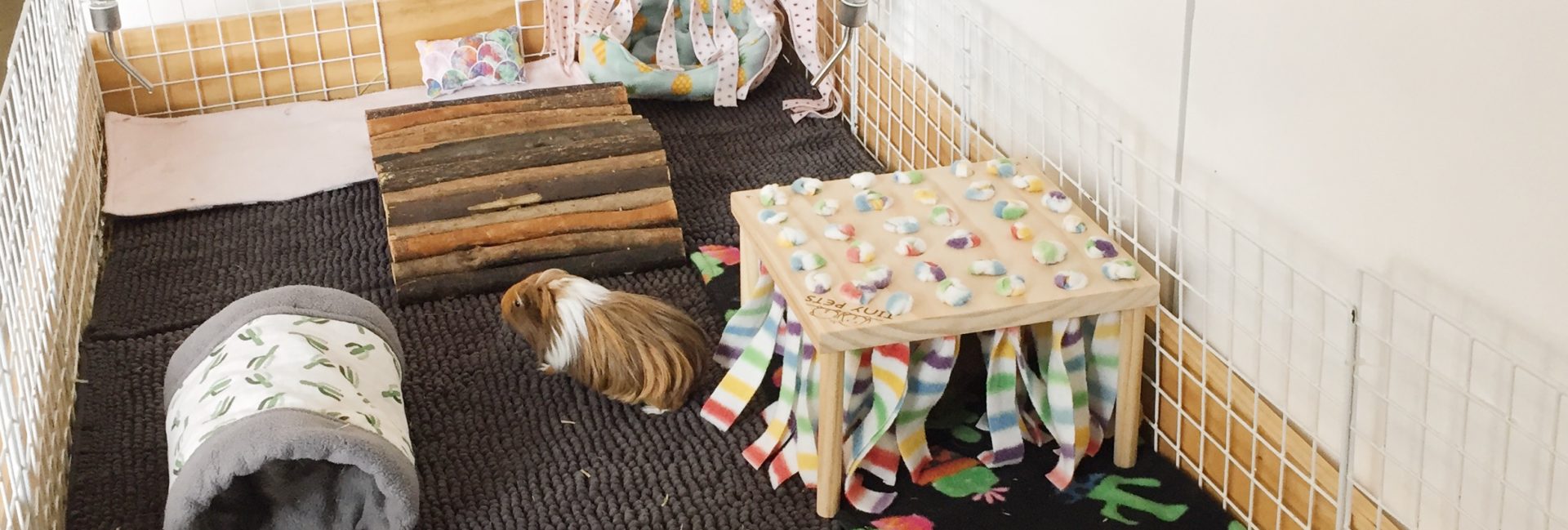 Olive and Ziggys guinea pig cage is handmade from plywood and a large wooden base. On the inside of the cage are three bath mats as liners, a wooden tunnel, a wooden hidey hut, some fleece cosy items and two large piles of hay and two water bottles.